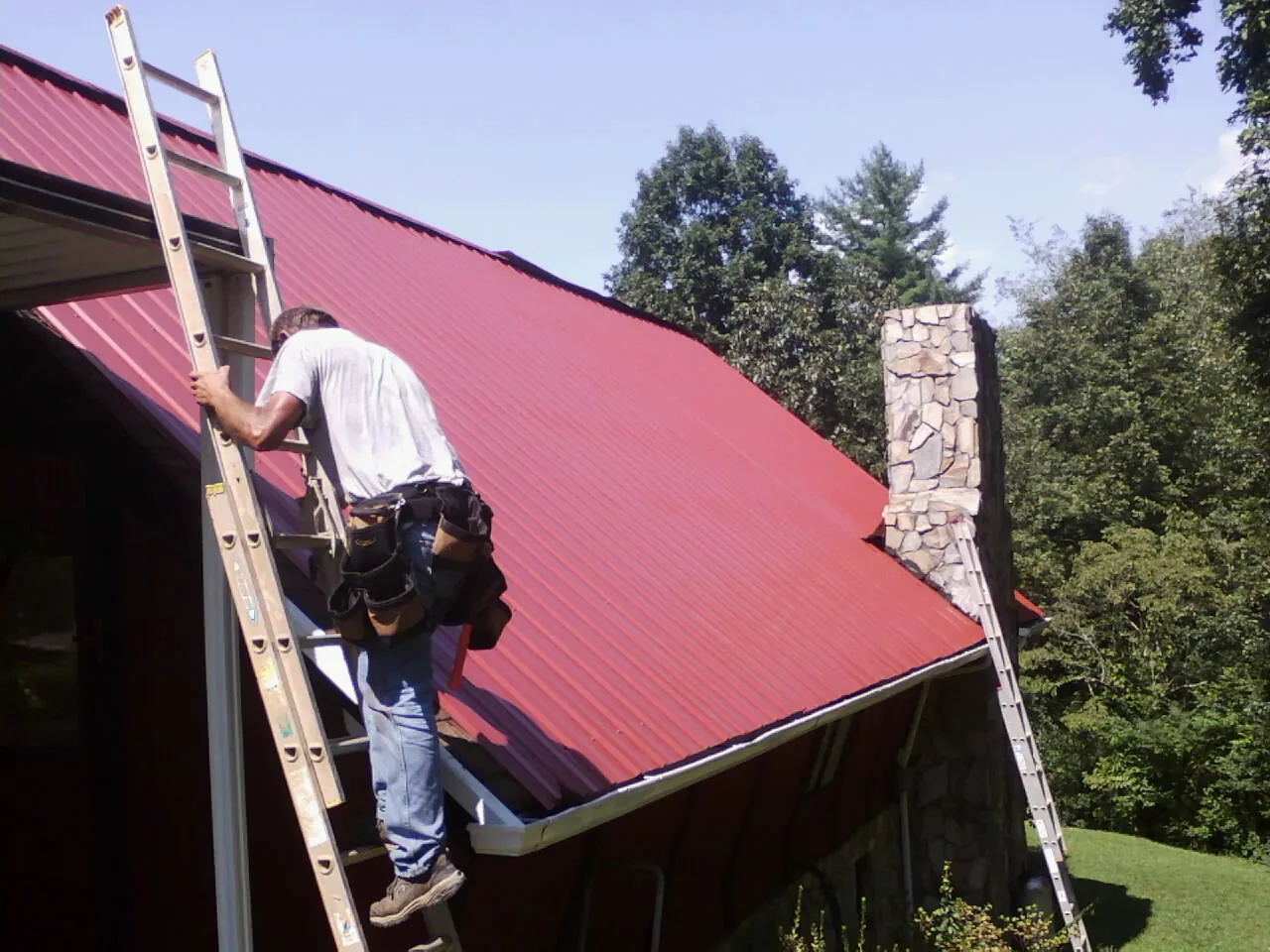 Roofer on a ladder beside a red metal roof and stone chimney — Soffit & Fascia Repair by Roofing Brothers in Fredericksburg